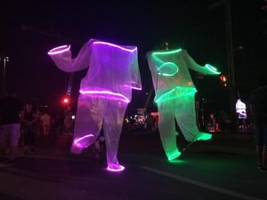Two Giant puppets dancing with people while bright LED strips border their head, arms and feet.
